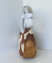 Load image into Gallery viewer, A cowhide shoulder bag with a brown and cream pattern, featuring a thick gray faux-leather shoulder strap wrapped in protective white foam and retail tags, set against a plain light gray background.