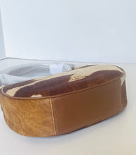 Load image into Gallery viewer, A small shoulder bag with a brown and beige cowhide pattern top, solid brown leather side panels, and a gold-tone logo plaque on top, resting on a white surface.