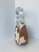 Load image into Gallery viewer, A cowhide shoulder bag with brown and cream patches, wrapped in protective white plastic stretch wrap on its handles, standing in a white studio corner.