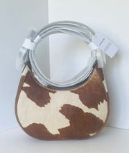 Load image into Gallery viewer, A small crescent-shaped shoulder bag with a brown and cream cowhide pattern, featuring a clear plastic-wrapped circular handle and a white hangtag.