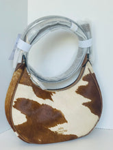 Load image into Gallery viewer, A cowhide shoulder bag with a brown and cream pattern, wrapped in protective plastic and held by white straps, with a small gold Coach logo plaque at the bottom center.