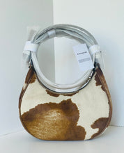 Load image into Gallery viewer, A Coach shoulder bag with a brown and cream cowhide pattern, featuring a light gray faux-leather handle wrapped in protective plastic and a white COACH brand tag.