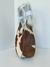 Load image into Gallery viewer, A cowhide shoulder bag with brown and white patches, featuring a curved handle wrapped in protective white paper and tape, set against a plain white background.