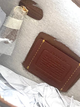 Load image into Gallery viewer, A close-up of a brown leather Coach patch with gold-tone stitching, placed on a white textured surface with crumpled white tissue paper and a small clear vial nearby.