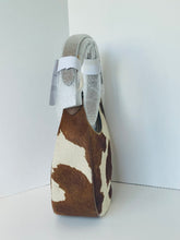 Load image into Gallery viewer, A cowhide shoulder bag with a brown and cream pattern, featuring a light gray fabric lining and white protective wrapping around the handle, set against a plain white background.