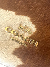 Load image into Gallery viewer, Gold-tone Coach New York logo charm and lettering on a brown and white cowhide surface.