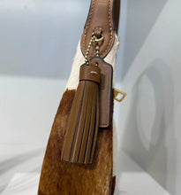 Load image into Gallery viewer, A close-up of a handbag featuring a brown leather tassel charm with a small gold-tone logo plaque, attached to a strap over a brown and white animal-print body.