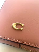 Load image into Gallery viewer, A close-up of a blush pink leather wallet featuring a signature gold-tone 'C' logo on the front flap and a gold zipper along the bottom edge.
