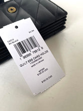 Load image into Gallery viewer, A black quilted wallet with a gold-tone snap button, shown with a white product tag reading 'Quilt ESS Card Holder' and other item details. The wallet is photographed on a light off-white surface.