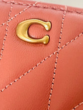 Load image into Gallery viewer, Close-up of a quilted coral pink leather item featuring a polished gold-tone 'C' logo on a white background.
