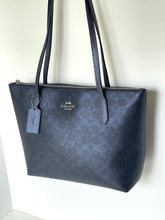 Load image into Gallery viewer, A dark navy blue Coach tote bag with a textured signature monogram pattern, featuring dual shoulder straps, a top zipper, and a hanging leather tag against a plain white wall.