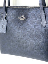 Load image into Gallery viewer, A close-up of a Coach handbag in textured navy blue leather, showing the silver-tone Coach logo on the front, a matching hanging luggage tag, and the top zipper.