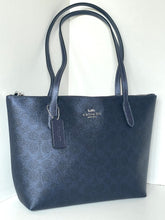 Load image into Gallery viewer, A dark navy blue pebbled leather tote bag with a subtle signature monogram pattern, featuring dual top handles, a silver-tone zipper, and a hanging brand tag. The Coach logo is visible on the front.