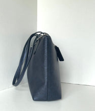 Load image into Gallery viewer, A dark navy blue textured leather shoulder bag with a matching strap and a small hanging tag, standing upright in a minimalist white corner setting.