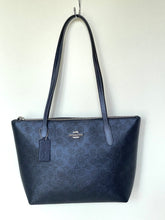 Load image into Gallery viewer, A Coach handbag in navy blue textured leather, hanging from a metal hook against a plain off-white wall. The bag features the Coach logo in silver-tone metal on the front and has two shoulder straps.