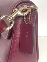 Load image into Gallery viewer, Close-up of a burgundy pebbled leather handbag with gold-tone hardware, a matching leather hangtag, and the embossed Coach logo.