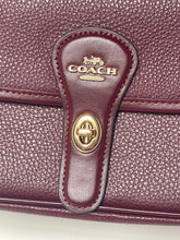 Load image into Gallery viewer, Close-up of a Coach handbag in pebbled burgundy leather, featuring a matching leather strap closure with gold-tone hardware and the embossed Coach logo.