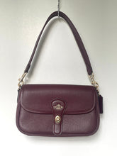 Load image into Gallery viewer, A Coach shoulder bag in dark burgundy pebbled leather, hanging from a metal hook against a light gray wall. The bag features a front flap with a gold-tone Coach logo and turn-lock closure, gold-tone hardware, and a matching burgundy leather strap.