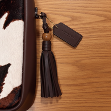 Load image into Gallery viewer, Brown leather tassel charm with a tag on a textured surface