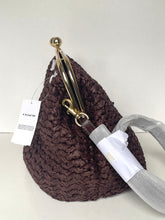 Load image into Gallery viewer, A Coach woven straw handbag in dark brown, featuring a gold-tone kiss-lock clasp and a white hangtag. The bag is shown with its protective white paper wrapping on the strap still attached.