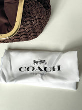 Load image into Gallery viewer, A Coach New York branded white dust bag with the black logo, placed on a light surface below a brown woven handbag with a gold-tone frame.