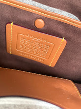 Load image into Gallery viewer, Close-up of the interior of a Coach bag showing a tan leather embossed brand patch on a brown fabric lining with yellow contrast stitching.