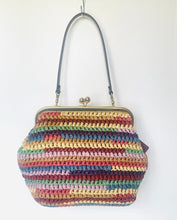 Load image into Gallery viewer, A handmade crocheted handbag with horizontal stripes of multiple colors including yellow, green, blue, red, and pink, featuring a dark gray shoulder strap and a gold-tone kiss-lock clasp, hanging against a plain white wall.