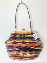 Load image into Gallery viewer, A multicolor crocheted handbag with a black shoulder strap and gold-tone metal frame, hanging against a plain white wall.