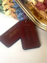 Load image into Gallery viewer, Two dark brown leather Coach key fobs on a white surface, with one fob showing the embossed Coach logo and the other showing gold-foiled text reading 'THE KEYBOARD FRAME BAG'.