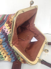 Load image into Gallery viewer, An open multicolor woven straw handbag with a gold-tone kiss-lock frame and a brown fabric lining, shown on a white background.