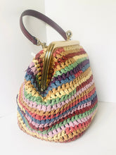 Load image into Gallery viewer, A handmade woven straw handbag with horizontal stripes of multiple colors, featuring a dark brown leather handle and a gold-tone metal kiss-lock clasp.
