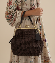 Load image into Gallery viewer, A woman wearing a bohemian floral print maxi dress and carrying a dark brown woven handbag with a gold-tone kiss-lock frame, standing in a modern living room setting.