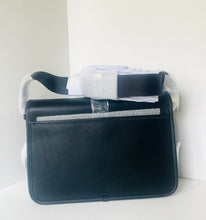 Load image into Gallery viewer, A black leather shoulder bag with a silver-tone metal bar detail, resting on a white surface against a white wall. The bag is still partially wrapped in protective white paper and plastic film.