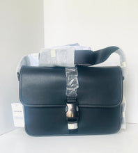 Load image into Gallery viewer, A Coach black leather messenger bag with a silver-tone turn-lock clasp, still wrapped in protective plastic and accompanied by its hangtag, set against a plain white background.