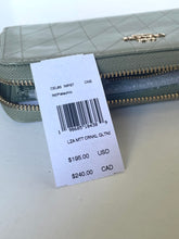 Load image into Gallery viewer, A quilted olive green wallet with gold-tone hardware and a white retail price tag attached, photographed on a light neutral surface.