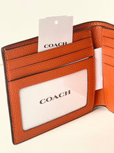 Load image into Gallery viewer, An open Coach bifold wallet in textured cognac brown leather, showing card slots, a clear ID window with the Coach logo, and a white hangtag.