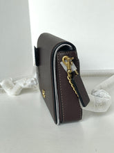 Load image into Gallery viewer, A dark brown pebbled leather crossbody bag with gold-tone hardware, shown standing upright on a white surface with protective white paper wrapping around its strap.