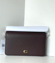 Load image into Gallery viewer, A dark burgundy pebbled leather crossbody bag with a gold-tone 'C' logo on the front, resting on a white surface with its white dust bag partially covering the top.