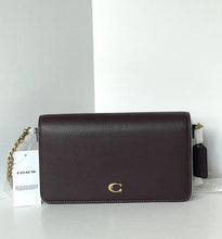 Load image into Gallery viewer, A Coach crossbody bag in dark burgundy pebbled leather with a gold-tone C logo on the front and a gold chain strap, shown with its hangtag on a white surface against a white background.