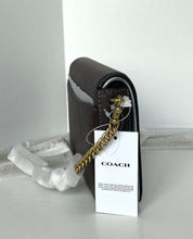 Load image into Gallery viewer, A Coach shoulder bag in dark brown textured leather with a gold chain strap and a white hangtag, resting on a light gray surface with white protective wrapping paper.