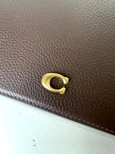 Load image into Gallery viewer, Close-up of a brown pebbled leather clutch or wallet featuring a polished gold-tone Coach 'C' logo clasp, resting on a light gray surface.