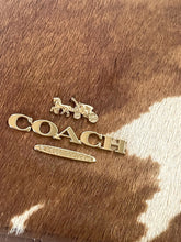 Load image into Gallery viewer, Gold Coach logo on a brown leather surface