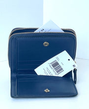 Load image into Gallery viewer, A dark navy blue leather zip-around wallet shown open, with interior card slots, a gold-tone snap button, zipper, and a white retail hangtag attached.