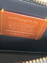 Load image into Gallery viewer, Close-up of a Coach interior label made of brown leather with gold stitching, showing the embossed COACH logo and text on a dark navy or black fabric surface.