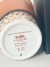 Load image into Gallery viewer, Close-up of the bottom of a Coach ceramic cup or tumbler, showing the white base printed with the Coach horse logo and care instructions, with part of the patterned rim and a dark box visible at the right edge.