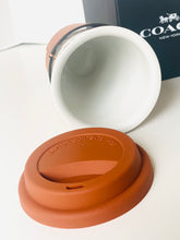 Load image into Gallery viewer, A ceramic mug with a terracotta-colored silicone lid and a black exterior band, shown with its lid placed in front and a Coach New York box in the background.