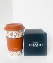 Load image into Gallery viewer, A Coach branded reusable coffee cup with a brown lid and sleeve, next to a black Coach New York gift box on a white background.