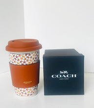 Load image into Gallery viewer, A Coach branded reusable coffee cup with a brown silicone sleeve and lid, standing next to its dark navy blue branded box on a white surface.