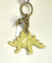 Load image into Gallery viewer, A pale yellow dinosaur keychain charm with a stegosaurus design, attached to a gold-tone split ring and keychain hardware, shown against a plain off-white background.
