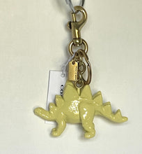 Load image into Gallery viewer, A Coach keychain featuring a pale yellow stegosaurus charm with gold-tone hardware, a silver split ring, and a white brand tag.
